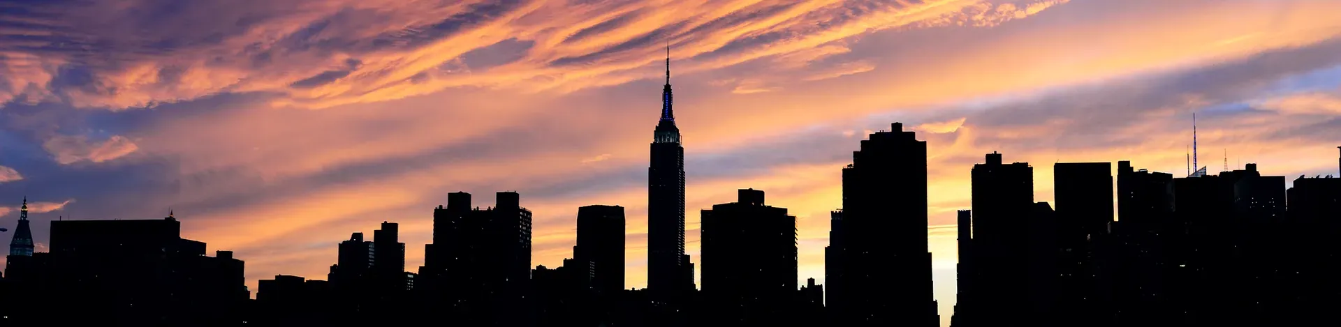 A city skyline in dark silhouette against a sunset sky streaked with pink and orange cloud
