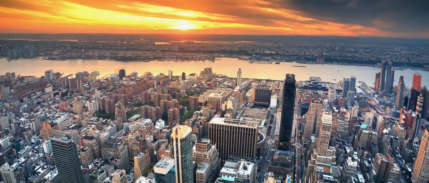 A city seen from above at sunset, the river and the far shore glowing under orange cloud